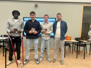 Four men pose indoors, holding awards, with a screen and snacks in the background.